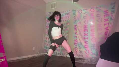 Media: Video of a slender, pale-skinned woman with black hair and glasses, wearing a black crop top, black shorts, and knee-high socks, dancing in a dimly lit room with a pink and blue graffiti wall.