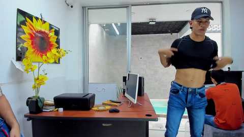 Media: Video of a fit, muscular man with a toned stomach, wearing a black Yankees cap and a black crop top, holding a PlayStation controller, standing in a modern, minimalist office with a large sunflower painting, a desk, and a pool view.