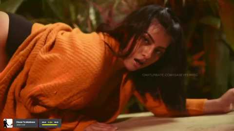 Media: Video of a woman with dark hair, wearing an orange knit sweater, on all fours, looking surprised. Background features blurred greenery.