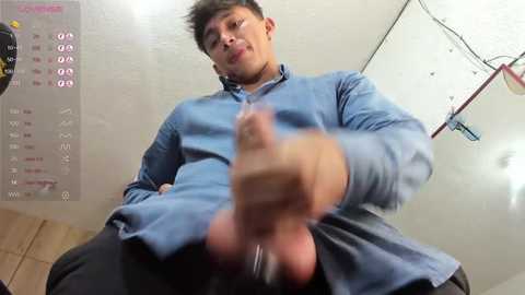 Media: A video of a man with a large erect penis, wearing a blue shirt and black pants, taken from a low angle. Background shows a white wall with a calendar and a clock.