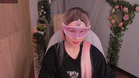 Media: Video of a young woman with long, straight, pastel-pink hair, wearing a purple face mask, black hoodie, and pink backpack, seated in a room decorated with floral wreaths and greenery.