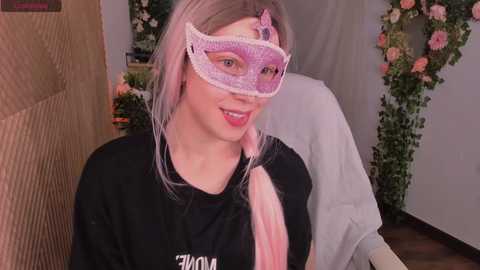 Media: Video of a young, fair-skinned woman with light pink hair, wearing a black shirt and a translucent, pink masquerade mask with a bow. Background includes floral decorations and a beige wall.