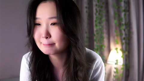 Media: Video of a serene, light-skinned Asian woman with long black hair, wearing a white top, smiling softly. She is indoors, with a lit table lamp and green curtains in the background.