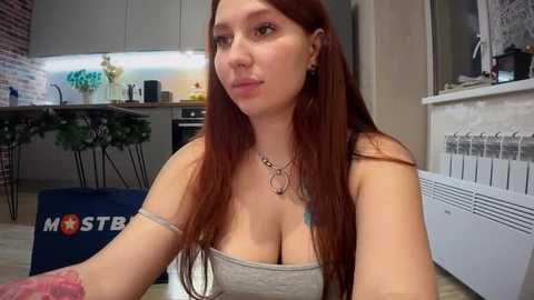 Media: A video of a young, light-skinned woman with long, straight red hair, wearing a gray tube top, sitting in a modern kitchen. She has a tattoo on her left arm.