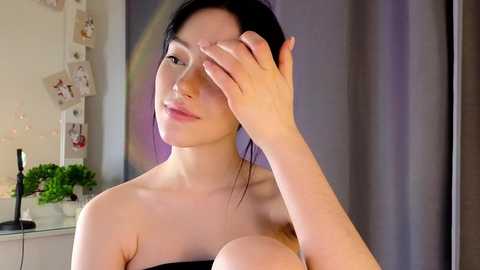 Media: Video of a young, slender Asian woman with pale skin, black hair, and light makeup, wearing a strapless black top, covering her left breast. She touches her hair with her right hand. Background includes a white wall, a potted plant, and a lamp.