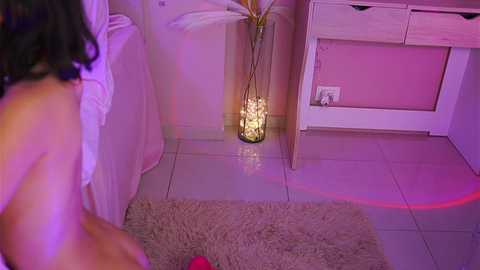 Media: Video of a nude woman with dark hair kneeling on a shaggy rug, beside a white bed with a lit candle stand and white furniture, bathed in pink and purple lighting.