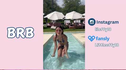 Media: Video of a young woman with long brown hair, wearing a black and white bikini, kneeling in a swimming pool, surrounded by white umbrellas and lounge chairs, set against a lush green background.