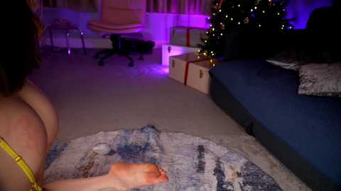 Media: Video of a woman with fair skin and brown hair, wearing a yellow bra, lying on a blue-patterned rug in a dimly lit living room with a Christmas tree and presents.