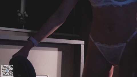 Media: A dimly-lit video of a slender, light-skinned woman wearing white lace lingerie, standing near a wooden desk with a black candle. Her left arm is extended, holding a black hat.