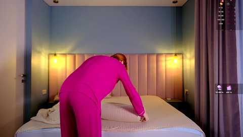 Media: Video of a person in a bright pink suit bending over a bed with white sheets, against a blue and beige wall, with a modern, minimalist room setup.