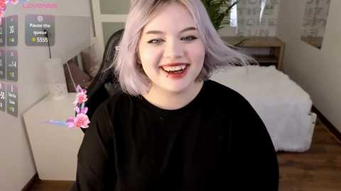 Media: Video of a smiling, light-skinned woman with shoulder-length, pastel pink hair, wearing a black top, sitting in a white chair in a modern, minimally decorated room.