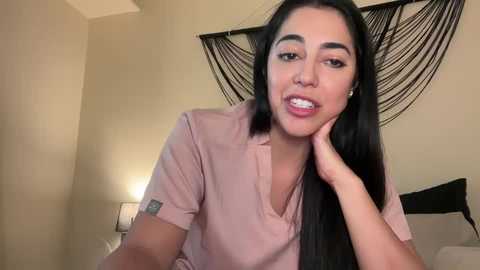 Media: Video of a Latina woman with long black hair, medium skin tone, wearing a pink shirt, smiling with her hand on her cheek, in a modern bedroom with beige walls and black decorative art.