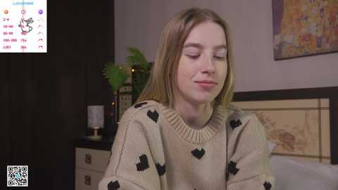 Media: A video of a young Caucasian woman with long blonde hair, wearing a beige sweater with black heart patterns, sitting on a bed, eyes closed in a serene pose.