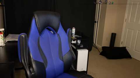 Media: Video of a modern gaming chair in vibrant blue and black, positioned in a dimly lit, cluttered room with a beige carpet, a white door, and scattered office supplies.