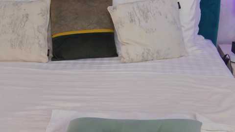 Media: Video of a neatly made bed with white linens, three pillows with various patterns, and a teal headboard. The pillows include a gray and yellow striped one, a white with gray pattern, and a solid white. The bedspread has a subtle texture.