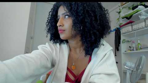 Media: Video of a young Black woman with a curly afro, wearing a white hoodie and red bra, seated indoors with a potted plant in the background.