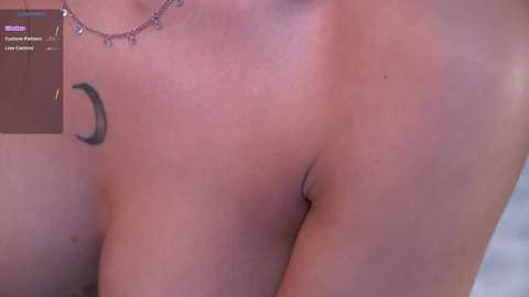 Video of a nude person with fair skin, showing a crescent moon tattoo on the left shoulder and a silver necklace, with blurred background.