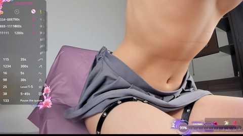 Media: A video shows a fair-skinned person with a flat stomach, wearing gray shorts pulled down, revealing black garter straps and panties. The background features a calendar, pink flowers, and a purple cushion.