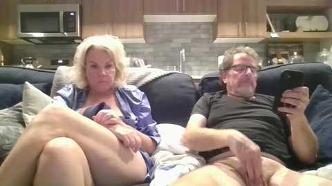 Media: Video of two middle-aged white people on a sofa, the woman with blonde hair in a blue robe, the man with glasses and dark hair in a black shirt, both nude below the waist.