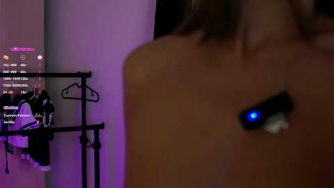 Media: Video: A blurred, topless woman with a glowing blue light on her neck stands in a dimly lit room with purple accents. A metal hanger with clothes hangs in the background.