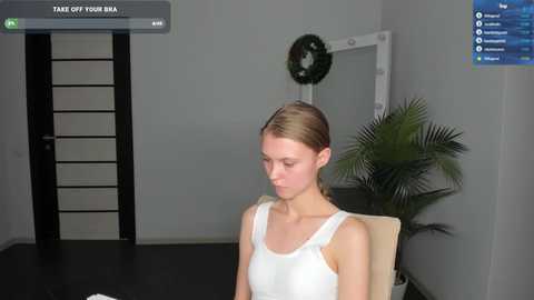 Media: A video of a young woman in a white tank top, sitting in an office chair, with a green plant and a black door in the background.