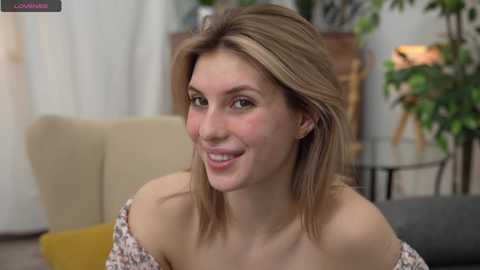 Media: Video of a smiling Caucasian woman with shoulder-length blonde hair, wearing a floral off-shoulder top, in a modern living room with greenery and beige furniture.