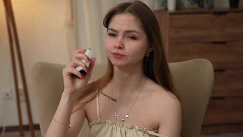 Media: A video of a young Caucasian woman with long brown hair, wearing a beige strapless top, holding a nasal spray. She sits in a beige armchair in a softly lit room with wooden furniture.