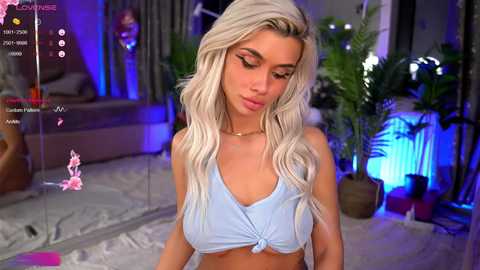 Media: Video of a light-skinned, blonde woman with long, wavy hair, wearing a light blue tied crop top, standing in a modern, dimly-lit room with potted plants, blue lighting, and a pink heart emoji on a screen.