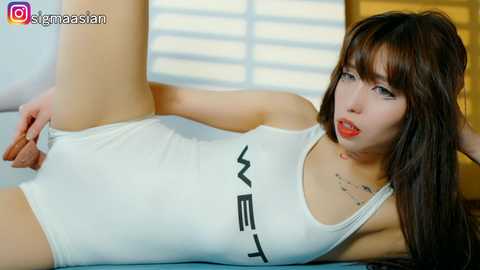 Video of a slim, fair-skinned Asian woman in a white, form-fitting bodysuit, lying on her back with legs spread, revealing her erect penis. She has long, straight brown hair and wears red lipstick.