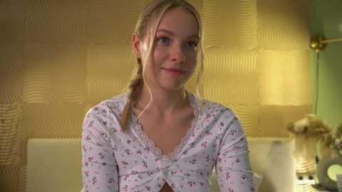 Media: Video of a smiling, light-skinned woman with blonde hair in braids, wearing a floral-patterned shirt, in a warmly lit, beige-textured room with a golden lamp.