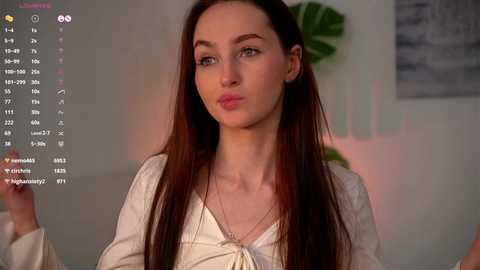 Media: Video of a young Caucasian woman with long, straight brown hair and fair skin, wearing a beige top, sitting indoors with a blurred background featuring a green plant and abstract artwork.