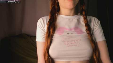 Video of a young woman with long red hair in pigtails, wearing a white crop top that reveals her medium-sized breasts and a pink heart design. Background includes a dimly lit bedroom with a bed.