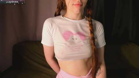 Media: A video of a young woman with fair skin, long red braided hair, wearing a white cropped t-shirt with a pink bunny graphic and light pink shorts, standing indoors with dim lighting and a couch in the background.