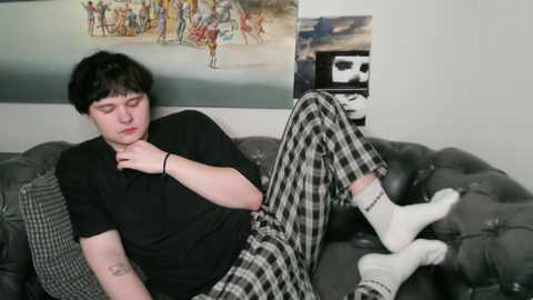 Media: Video of a young man with short black hair, wearing black shirt, white socks, and plaid pajama pants, lying on a gray couch, in a room with wall art.