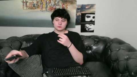 Media: Video of a young man with short black hair, wearing a black shirt, seated on a dark gray, tufted leather sofa. Background features a wall with colorful, abstract art and a black and white skull portrait.