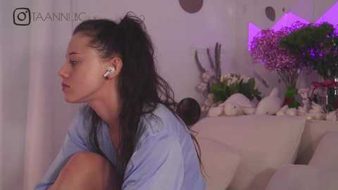 Media: Video of a young woman with long black hair in a ponytail, wearing a light blue shirt, sitting thoughtfully in a modern living room with white decor, purple ambient lighting, and floral arrangements.