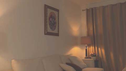 Media: A video of a dimly lit living room with beige walls, a white sofa, a framed circular painting, and a table lamp with a beige lampshade.