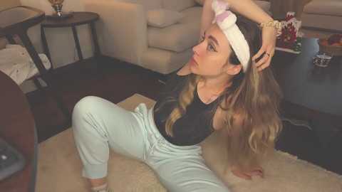 Media: Video of a blonde woman in a black tank top and light blue joggers, adjusting a white headband on a beige carpet in a living room with beige furniture.