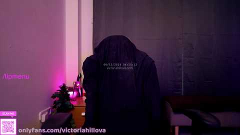 Media: Video of a person with long black hair, wearing a dark jacket, standing in a dimly lit room with purple and pink lighting. The background includes a wall, a chair, and a plant. Text overlays.