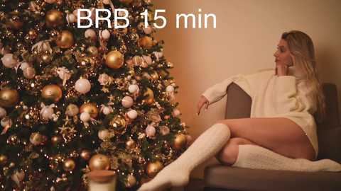 Media: Video of a blonde woman in a cozy white sweater and knee-high socks, sitting on a brown armchair, gazing at a glittering, gold-decorated Christmas tree in a warm, beige-lit room. Text reads \"BBR 15 min.\