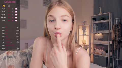 Media: A video of a young, light-skinned woman with blonde hair, wearing a white tank top, playfully covering her mouth with her finger in a bedroom with a bed, shelves, and a lamp.