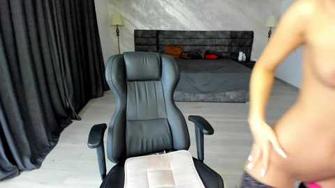 Media: Video of a modern bedroom featuring a black leather gaming chair, a grey bed with orange pillows, and a nude woman standing in the background.