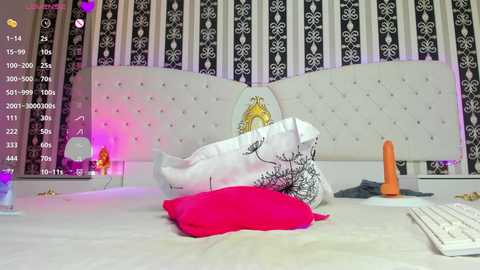 Media: Video of a modern bedroom with a white tufted headboard, black and white floral wallpaper, a white duvet with a red pillow, and an orange dildo on the bed.