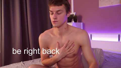 Media: Video of a young, shirtless man with light skin, short brown hair, and a toned physique, sitting on a bed. He covers his chest with one hand. Background includes a pink wall, gray curtains, and a plant. Text overlays: \"be right back.\