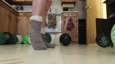 Media: Video of a person in grey, white-knit socks and a white tank top, standing on a tiled kitchen floor, surrounded by scattered green and black balloons.