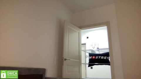 Media: Video of a simple, empty room with a partially open white door revealing a black t-shirt with white text. The room has beige walls and a grey couch in the corner.