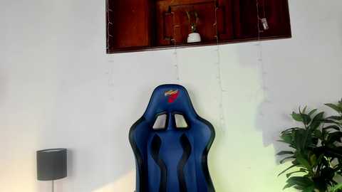 Media: Video of a blue gaming chair with ergonomic design, mounted on a white wall with a small wooden cabinet and plants, featuring a small window with fairy lights.