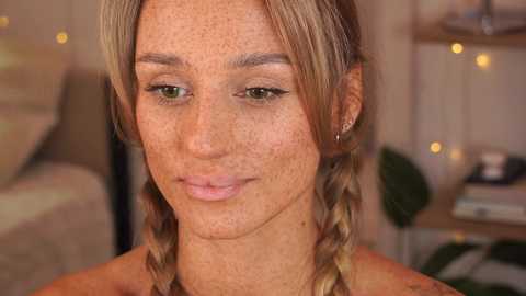 Media: Video of a fair-skinned woman with blonde braids, freckles, and green eyes, wearing minimal makeup. Background features a blurred, cozy indoor setting with warm lighting and green plants.