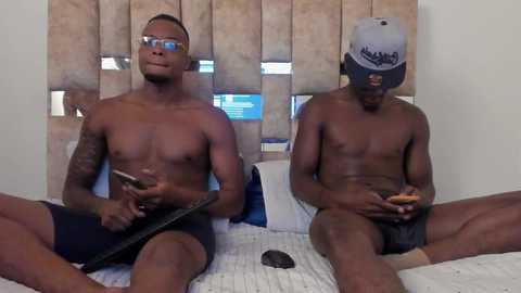 Media: Video of two shirtless, muscular Black men with tattoos, sitting on a bed with a beige headboard, holding smartphones, wearing sunglasses, and baseball caps.