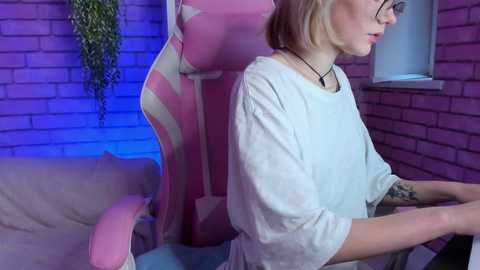 Media: Video of a slender, fair-skinned woman with shoulder-length blonde hair, wearing glasses and a white T-shirt, playing on a pink gaming chair in a brick-walled room with purple lighting and a hanging plant.